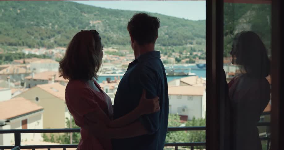 A man and woman excitedly embrace and kiss while looking out at a nearby bay from their balcony during the day