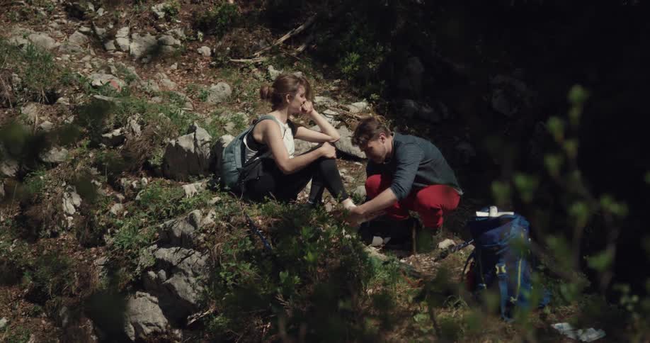 Male hiker helps wrap ankle of female hiker as she sit on slope in wilderness