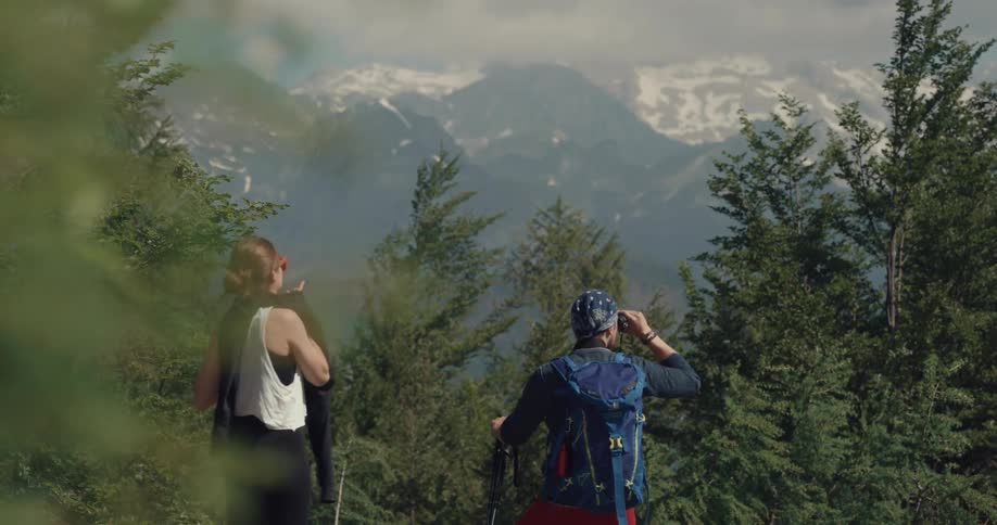 A man and woman stop during their hike and look at the mountains while the woman takes her heavier shirt off and ties it around her waist