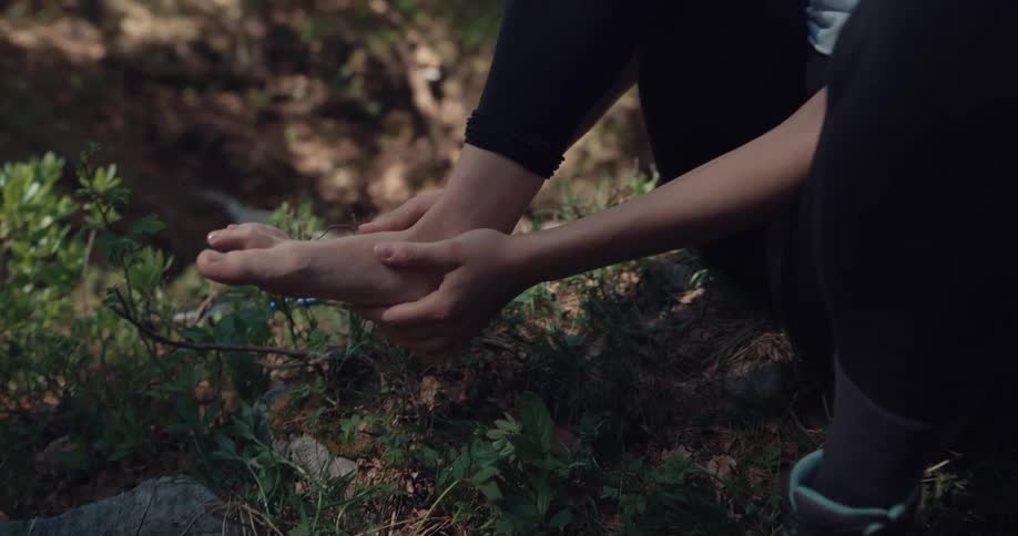 Woman sits on ground near woods grimacing and crying out in pain as she grasps ankle