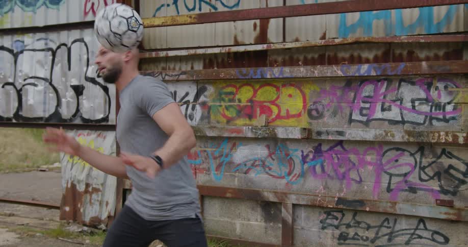 A man doing a trick with a soccer ball on his back with a wall with graffiti in the background