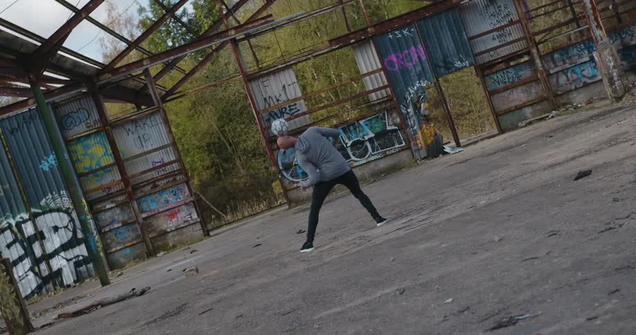 A man juggles a soccer ball using various parts of his body while standing in a dilapilated building during the day