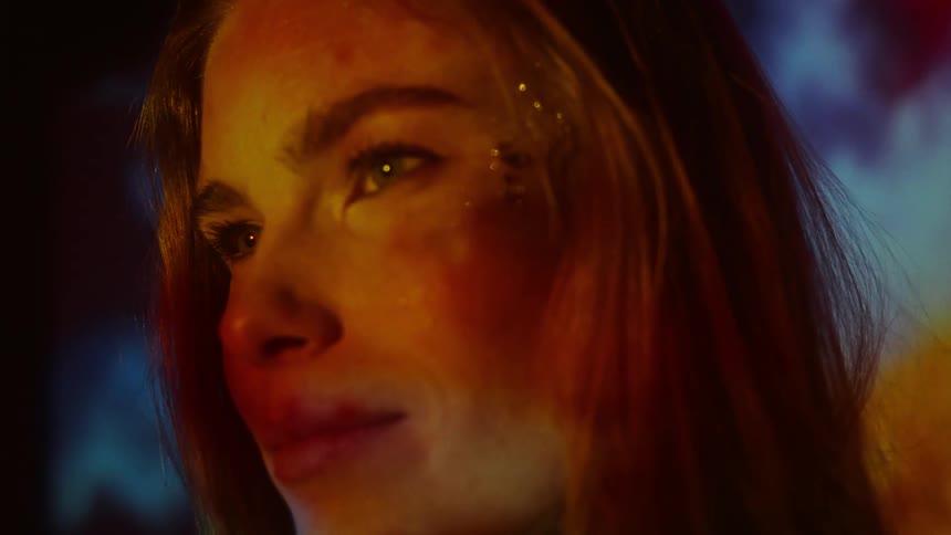 A woman stands in a dark room as colorful light is projected onto her face
