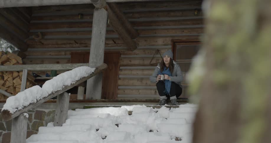 A woman walks out of a wood cabin and sits down