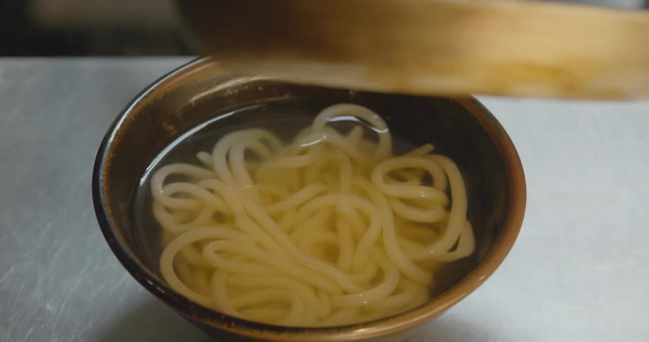 Bacon and eggs are poured from a skillet into a steaming bowl of noodles before the bowl is taken away