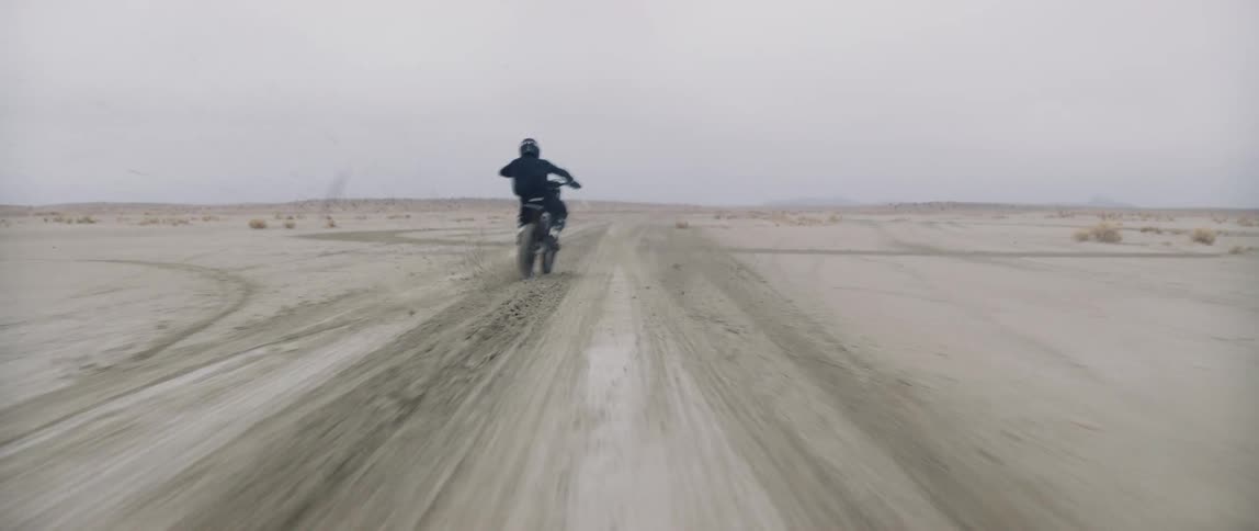 A person with a helmet riding a dirt bike on a desert landscape with the sky and clouds in the background