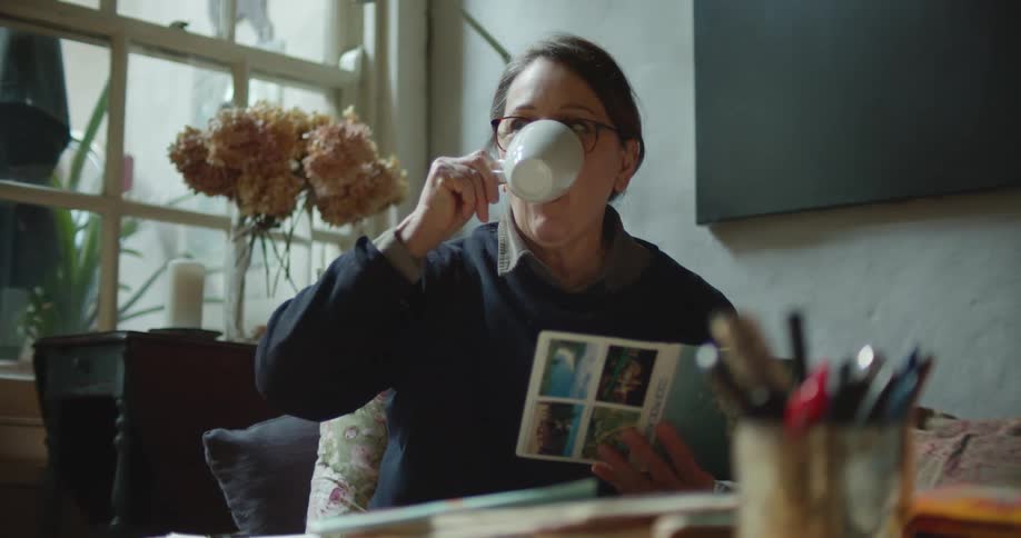 Woman wearing glasses drinks a beverage from a mug while looking at a brochure