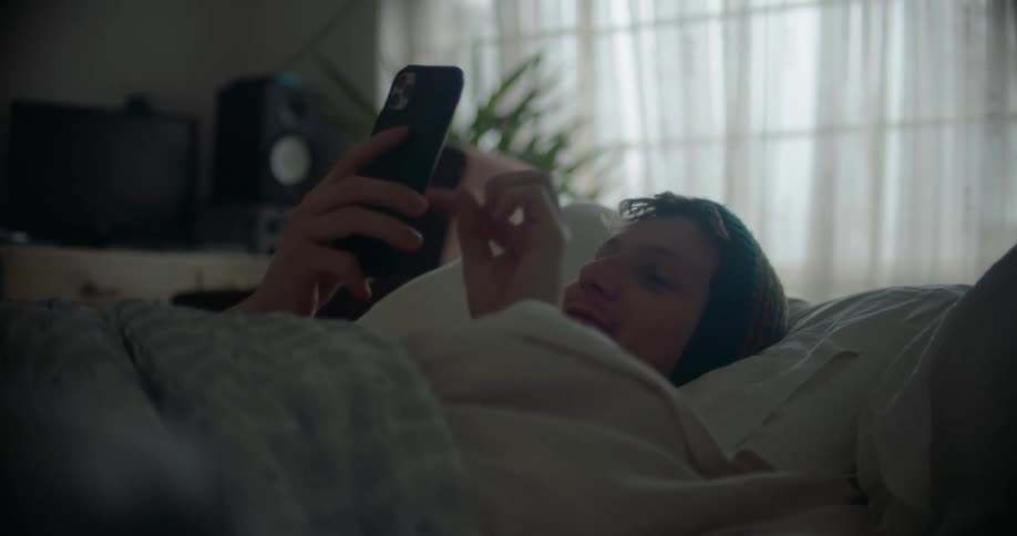 Man wearing knit hat lies in bed under covers using mobile phone and smiling