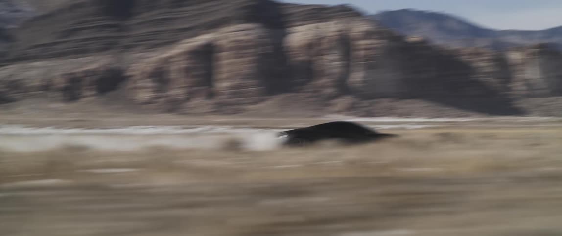 A car leaves a dust trail as it races on flat land pas brush and rock formations