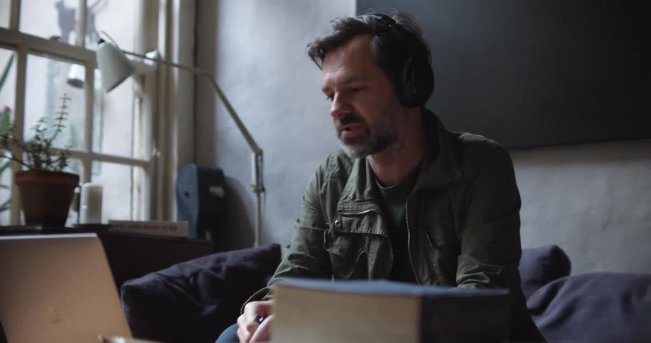 Man wearing headphones talks to someone on his laptop while he holds a pen in his right hand