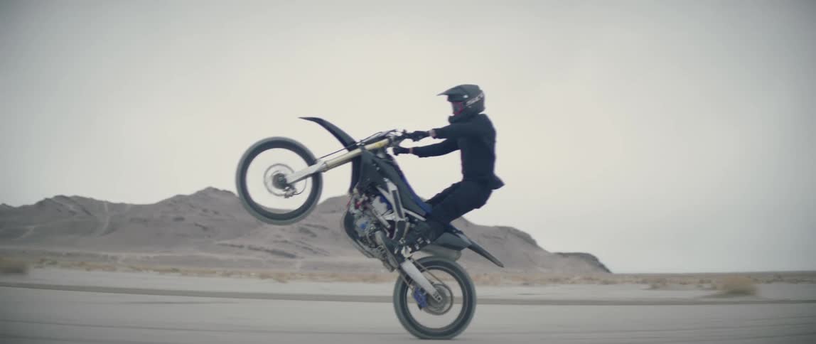 Man rides motorcycle during extended wheelie over dirt passing rocky hills
