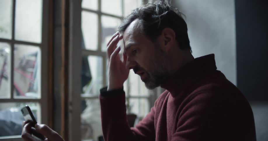 A man in a room talking on a smart phone with a wall and window in the background