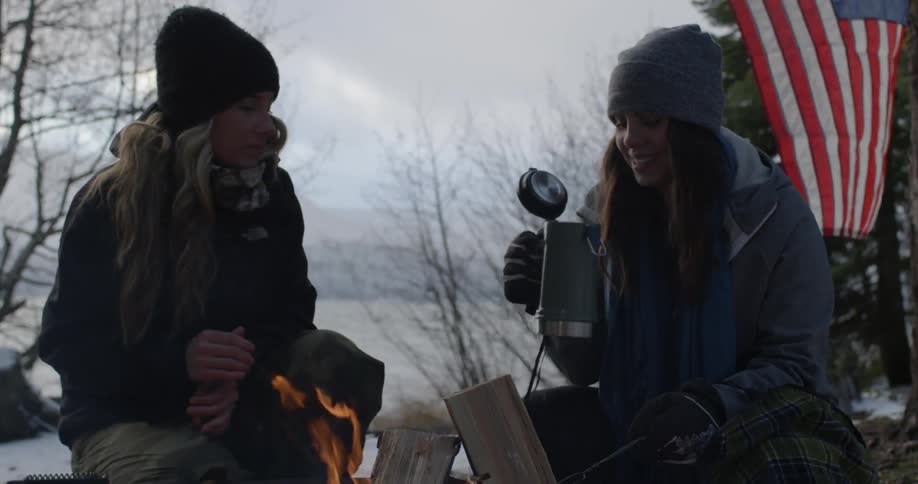 Two women drink from a thermos while sitting next to a fire on a winter day