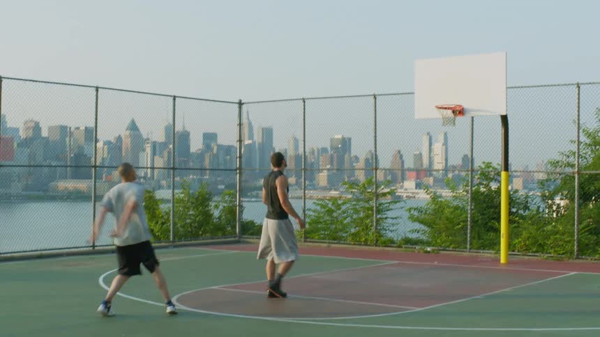 A man shooting a basketball while another guards him on a basketball court located next to a body of water in an urban area