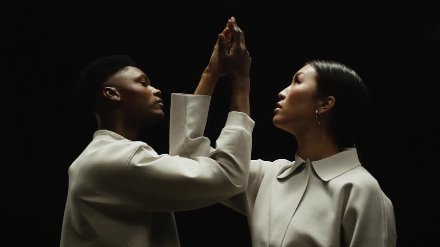 Black man and black woman stand facing each other with arms raised and their fingers touching
