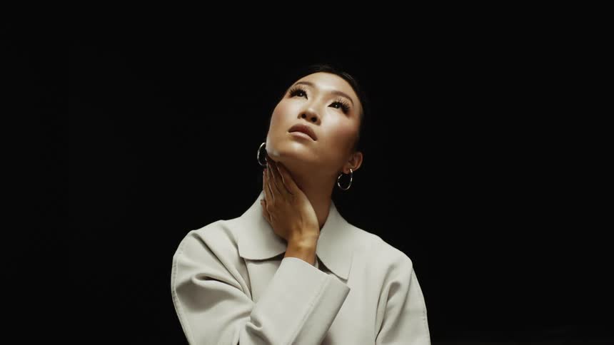 Woman wearing off white clothing holds hand against her neck as she looks upward against black background