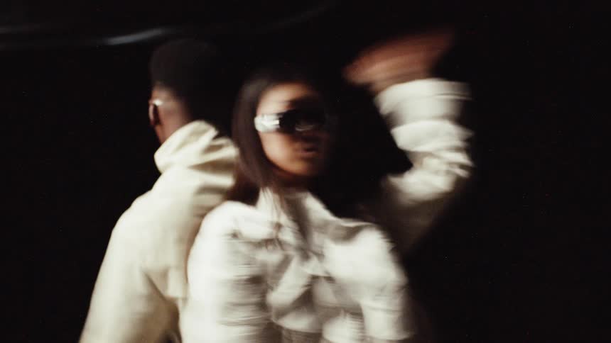 Man and woman wearing cream colored clothing and shades stand back to back moving their arms and dancing against black background