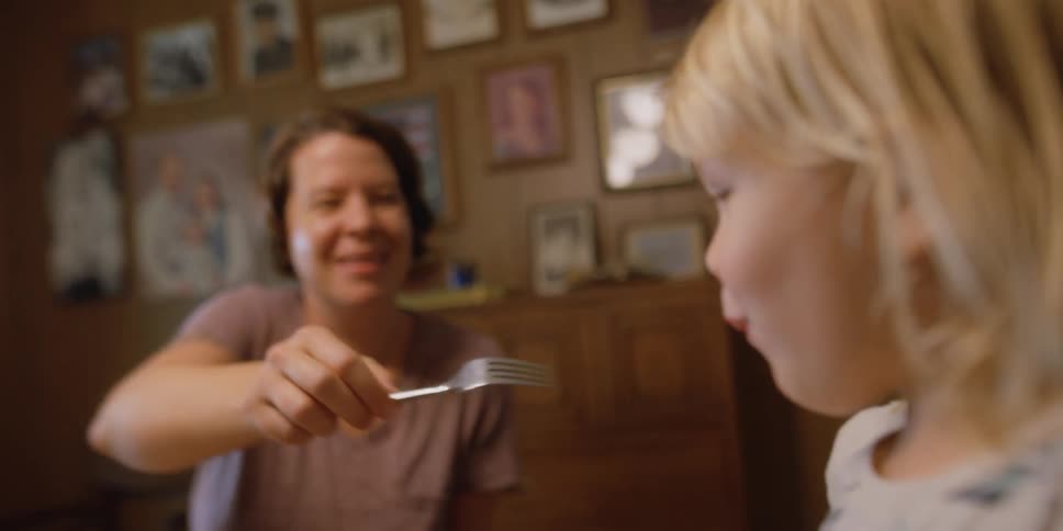 A woman in a room sitting at a table feeding a little girl with a fork