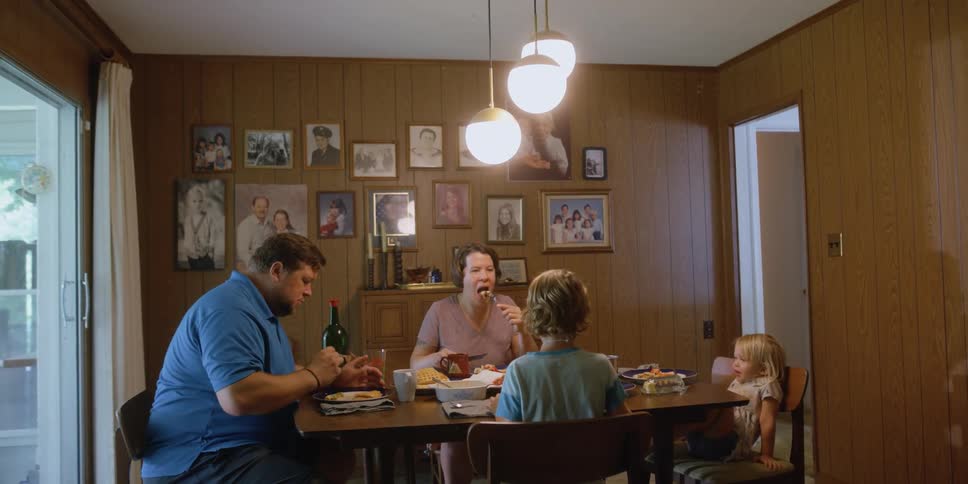 Family sits around dining table at eating and talking with framed family photographs on wooden paneled walls