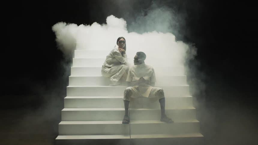 A man and a woman sit on a white staircase with smoke behind them