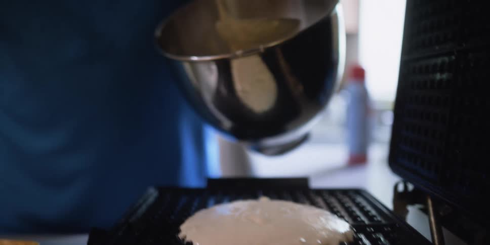 Man pouring batter from a bowl into a waffle maker on the kitchen counter