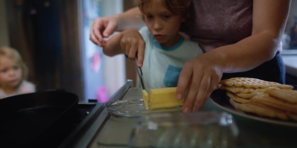 A young boy slices a piece of butter with the help of his mother behind him