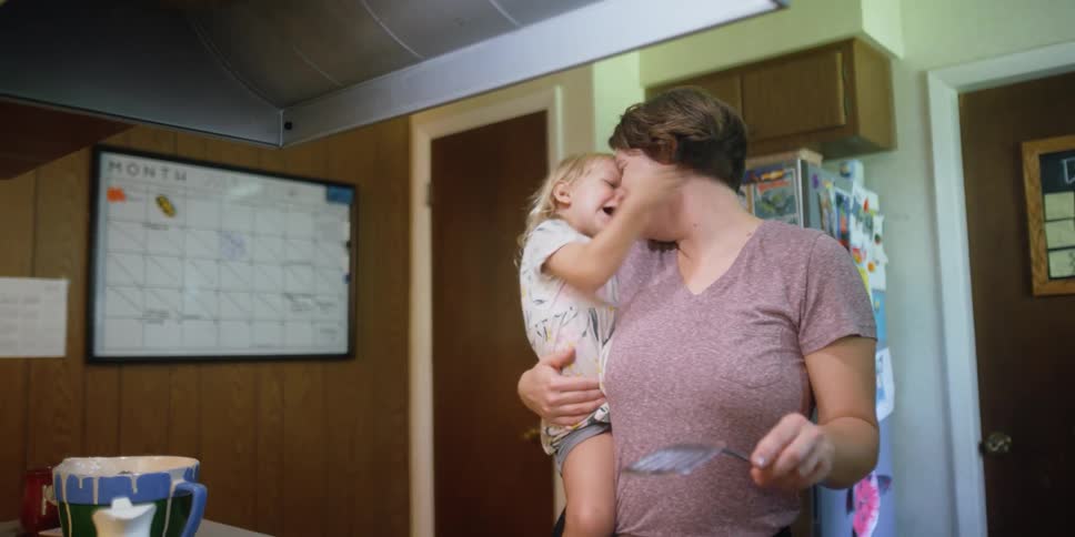 Woman holds a female toddler and they laugh and rub noses while the woman holds a spatula