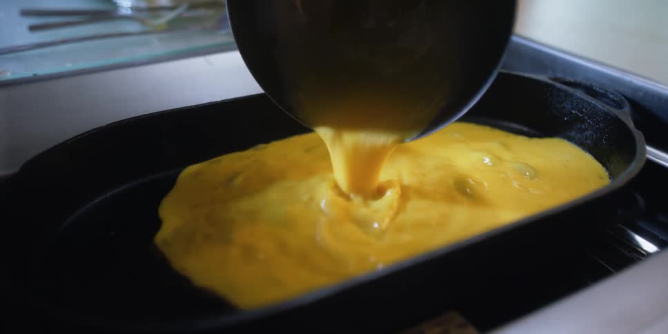 A woman in a kitchen pouring eggs from a bowl into a skillet on a stove