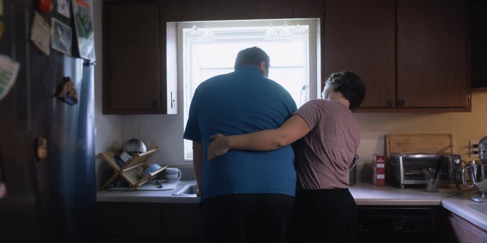 A woman greets a man washing dishes in the kitchen sink with a kiss and a back rub