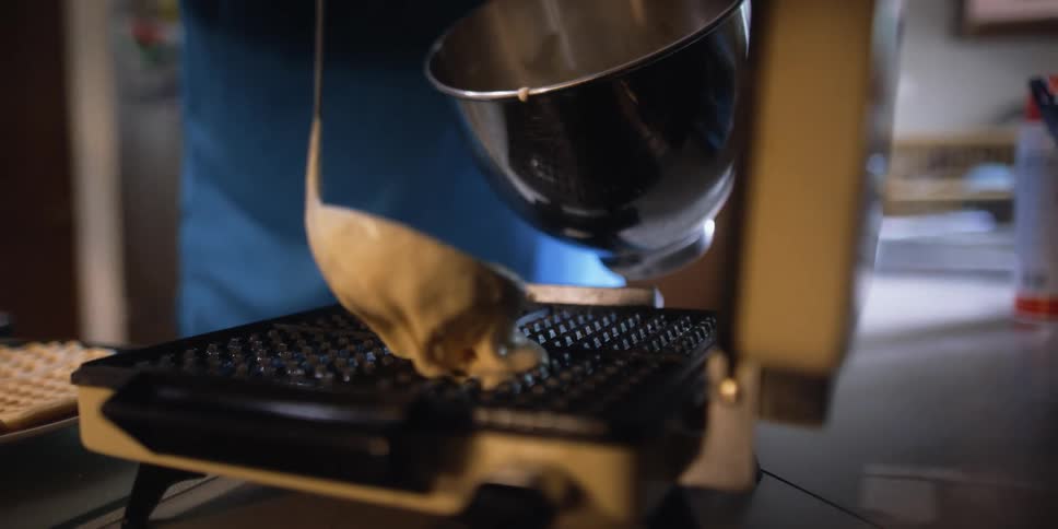 A man in a kitchen pouring pancake batter on a waffle iron with a woman in the background