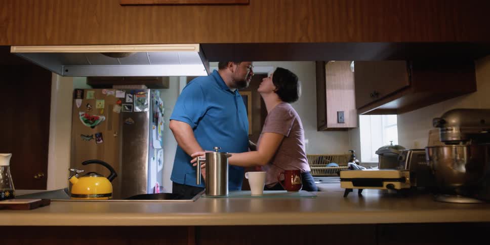 Woman standing in kitchen sets coffee mug down to greet husband with a kiss