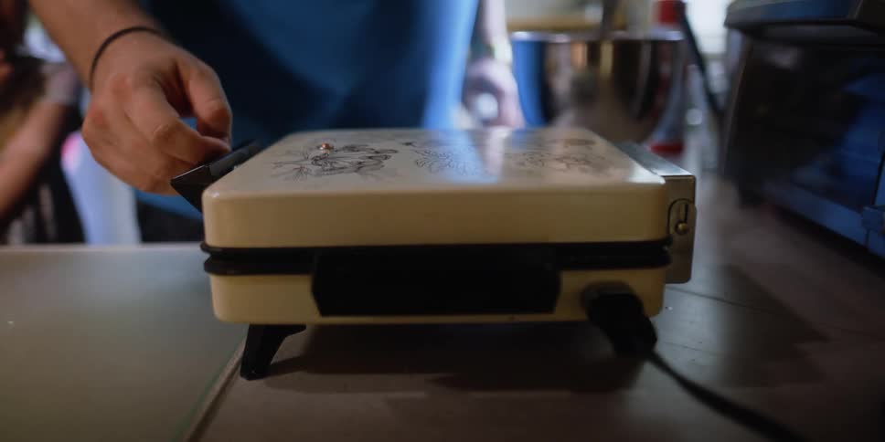 People are in a kitchen together while a man closes the top on a waffle maker