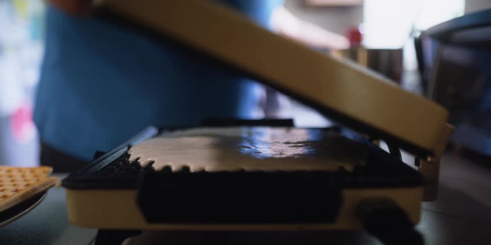 A person in a kitchen pouring pancake batter on a waffle iron with another person and a wall in the background