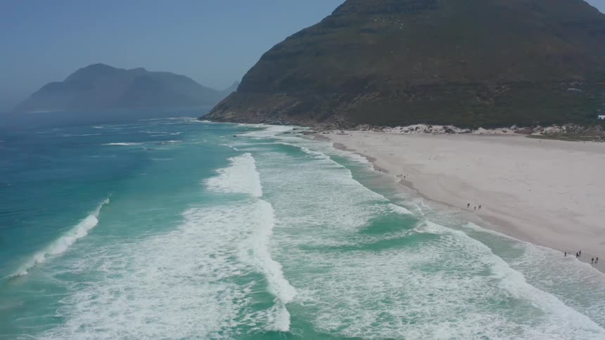 People are on the beach as waves roll over the sand with mountains in the distance