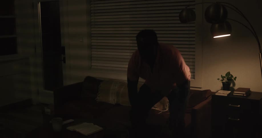 Man walking inside a dimly lit living room sits on a couch