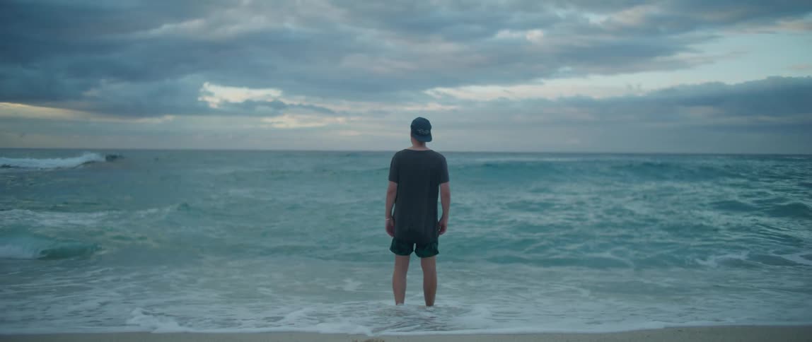 A man walks toward the ocean with his hands in his short pockets