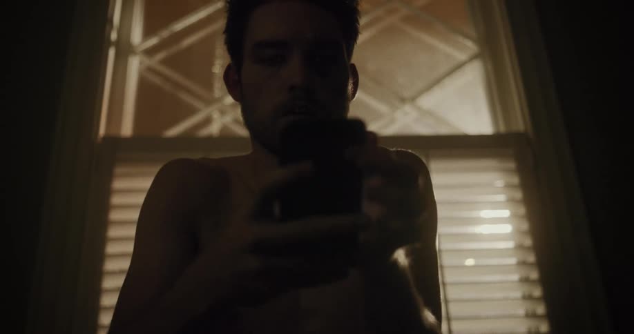 A man sitting down indoors in front of a window puts a gun down beside him and then picks up a phone and starts texting