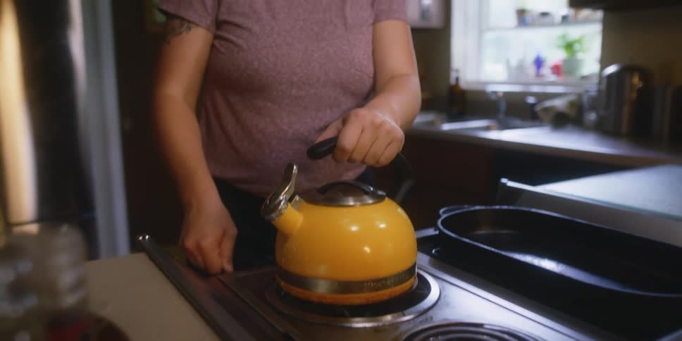 Woman places a yellow metallic whistling tea kettle over the kitchen range