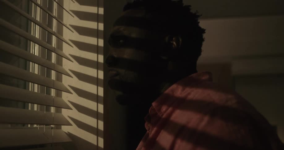 Fearful man inside a dimly lit room looks nervously out the window