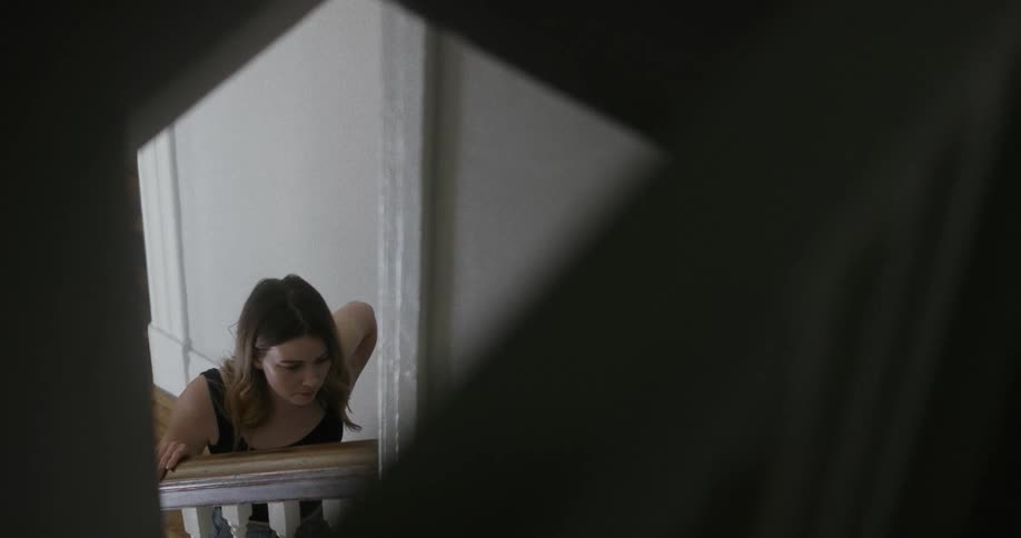 A woman in a room standing by a staircase and touching the banister with a wall in the background