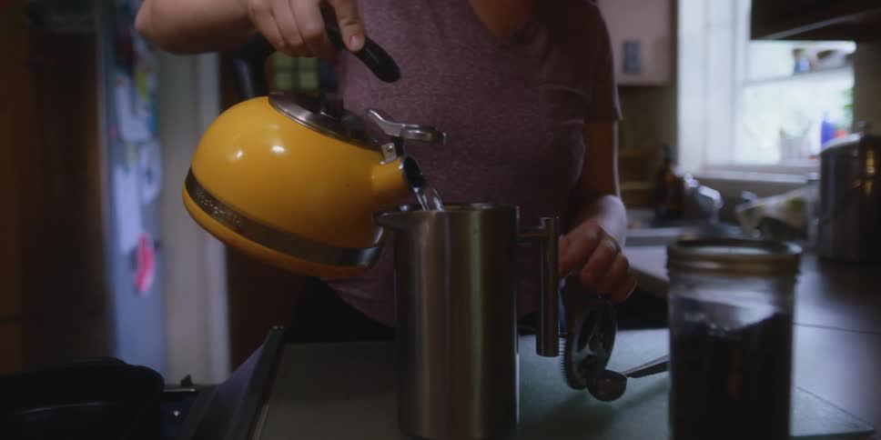 A woman in a kitchen pouring hot water from a kettle into a mug
