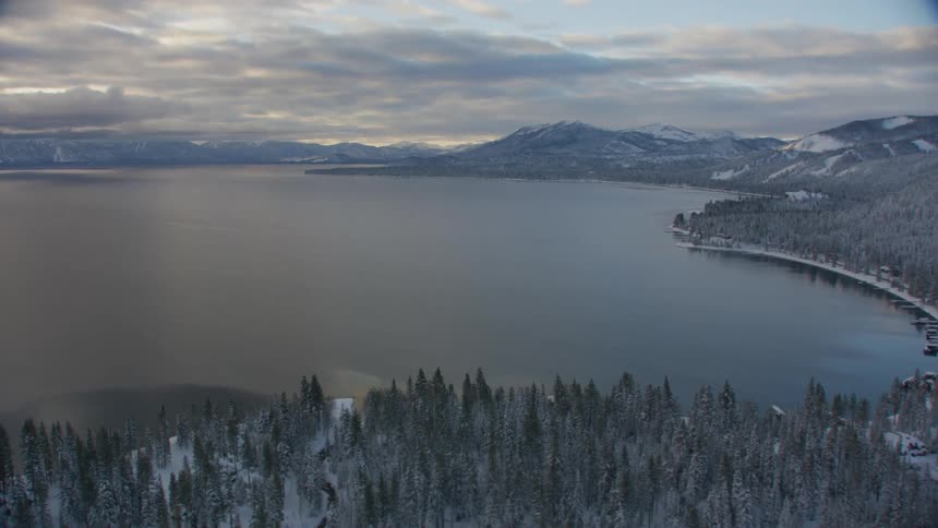 Snow covered forests and mountains surrounding a lake under a cloudy sky