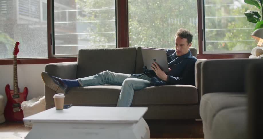 A man lays on a couch looking at an ipad tablet