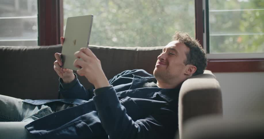 A man lays on a couch holding and reading an ipad