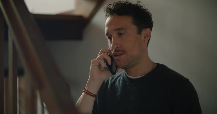 A man standing on a staircase talking on a smart phone with a banister in the foreground and a wall in the background