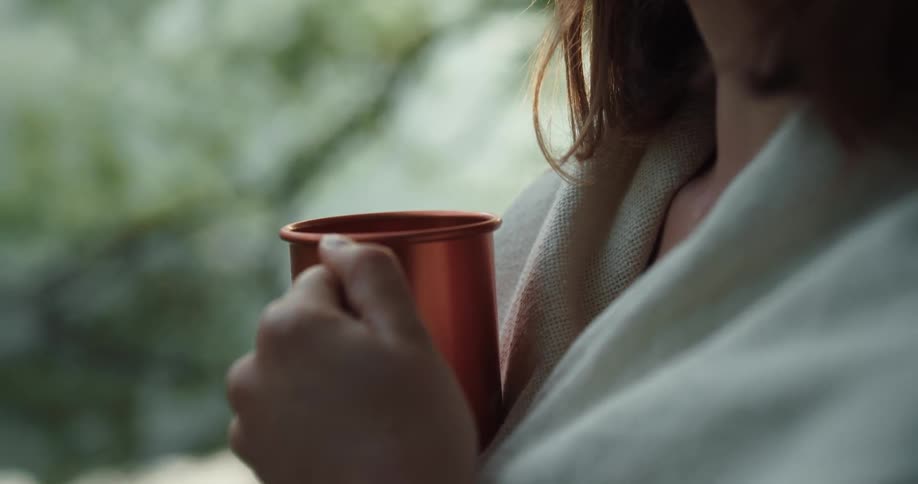 Woman with brown hair and beige shawl holds a copper cup in the forest