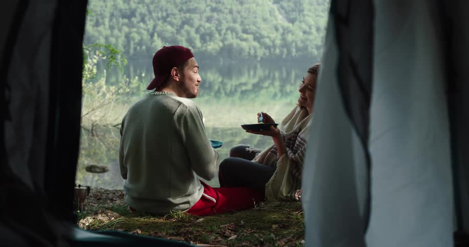 Two people sit in front of a tent and eat a meal together