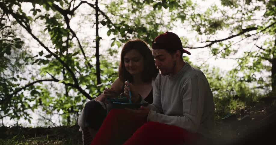 A pair of campers chat with one another while sitting side by side in a forest and eating