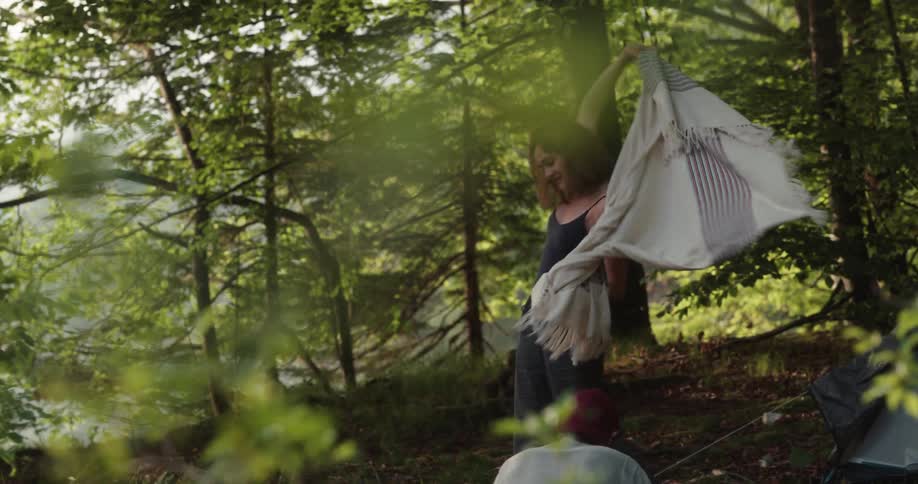 A woman climbs out of her tent in a forest and wraps herself in a shawl as a man approaches to hand her a cup of coffee