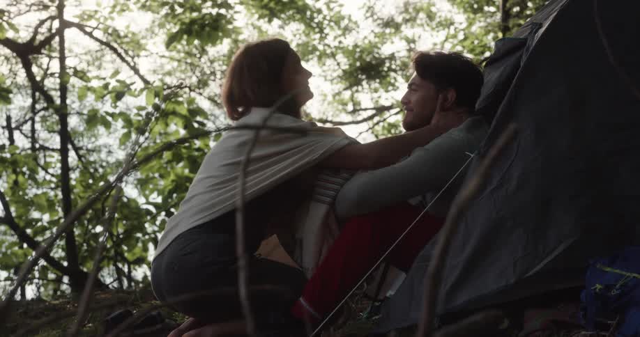 A woman walks up to her boyfriend and kisses him as he sits in the entrance of a tent propped up in the woods